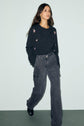 A woman stands against a pale background, wearing a dark gilet en laine with small motif fleuri embroidered flowers and loose-fitting gray cargo jeans. She has long brown hair and black shoes.