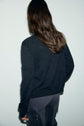A person with long brown hair stands with their back to the camera, wearing a dark sweater or gilet en laine and gray pants against a light background.