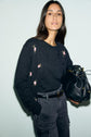 Woman with long brown hair wears a dark gilet en laine with pink motif fleuri embroidery, dark high-waisted jeans, and holds a black handbag against a pale background. She has a neutral expression and her hand is in her pocket.