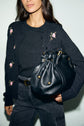 A woman wearing a dark gilet en laine à motif fleuri and black jeans holds a large black leather handbag with gold accents. The photo is cropped at the shoulders and thighs, focusing on her outfit and bag.