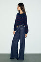 Woman standing against a light background, wearing a navy and rayures bordeaux marinière-style sweater with button details in soft laine mérinos, high-waisted wide-leg jeans, a black belt, and black shoes. She is looking to her left.