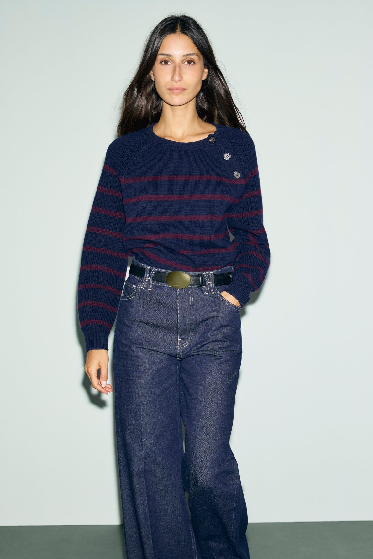 Woman with long brown hair wearing a navy and rayures bordeaux marinière-style sweater with button details in soft laine mérinos and high-waisted wide-leg jeans, standing against a plain light background.