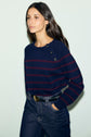 A woman wearing a navy blue marinière sweater with rayures bordeaux and button details on the shoulder, crafted from soft laine mérinos, is paired with high-waisted dark jeans and a black belt as she stands against a light background.