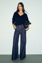 A woman stands against a plain background wearing a pull col V laine bleu marine, wide-leg dark jeans with a belt, and holding a black handbag. She has long brown hair and her hands are in her pockets.