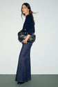 Woman standing against a plain wall, wearing a pull bleu marine sweater, wide-leg jeans, and black shoes, holding a large black handbag under her arm, looking slightly over her shoulder.