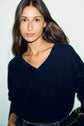 Woman with long brown hair wearing a pull bleu marine col V en laine and jeans, standing against a light background and holding a black handbag.