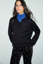 A woman with long brown hair wears a black pull-over over a blue polo laine mérinos à manches longues and dark pants with small blue floral patterns, standing against a light background.