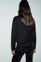 A person with long brown hair, wearing a black polo manches longues sweater and black pants with small blue flower patterns, stands facing away from the camera against a plain light background.