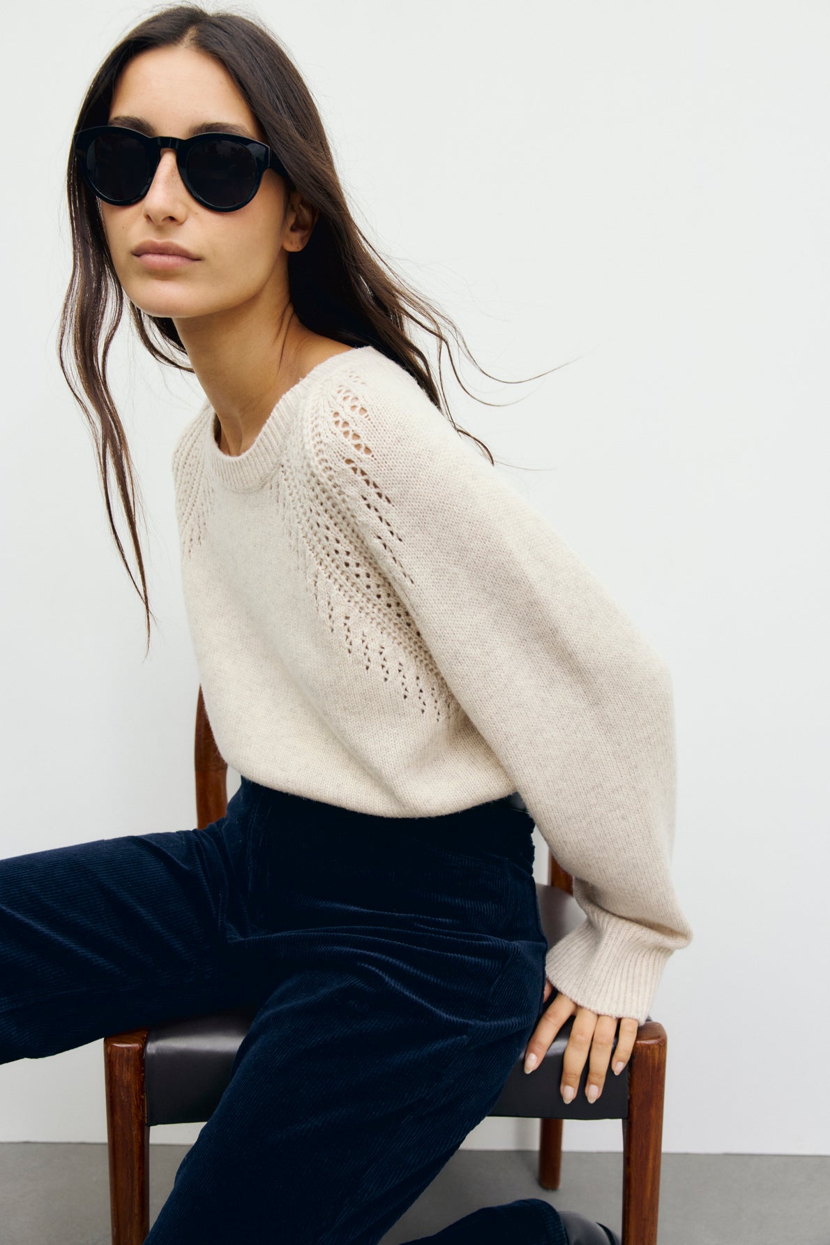 Woman wearing black sunglasses, a pull écru en laine, and dark blue corduroy pants sits on a wooden chair against a plain light-colored wall, looking slightly to the side.