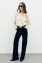 Woman wearing sunglasses, a pull écru en laine, black velvet wide-leg pants with a large belt buckle, and black shoes stands against a plain light background with hands in pockets.