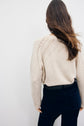 A person with long brown hair, seen from behind, wearing a pull écru en laine with a textured pattern and dark pants, standing against a plain light background.