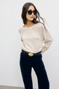 Woman in black sunglasses, beige pull en laine, and dark pants stands against a plain white background, with one hand in her pocket and her long hair loose. She wears a black belt with a large oval buckle.
