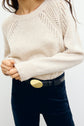 A person wearing a light beige pull écru en laine with a textured pattern, black pants, and a black belt with a large oval gold buckle. Their hand is holding the sweater at the waist.