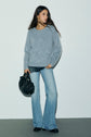 Woman in a light gray sweater crafted from pull bleu grisé cachemire brossé and flared blue jeans stands against a plain white wall, holding a black handbag and wearing casual black shoes.
