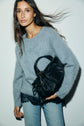 A woman with long brown hair wears a pull bleu grisé, lace-trimmed top, and light jeans, holding a black textured handbag against a plain light background.