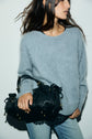 A woman in a light gray cachemire brossé sweater and light blue jeans holds a black handbag. Her hair is loose, and her face is partially turned away from the camera against a plain white background.