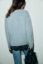 A person stands against a white wall, seen from behind, wearing a pull bleu grisé ultra doux in fuzzy light gray, paired with blue jeans and holding a black handbag. A black lace top peeks out from under the sweater.