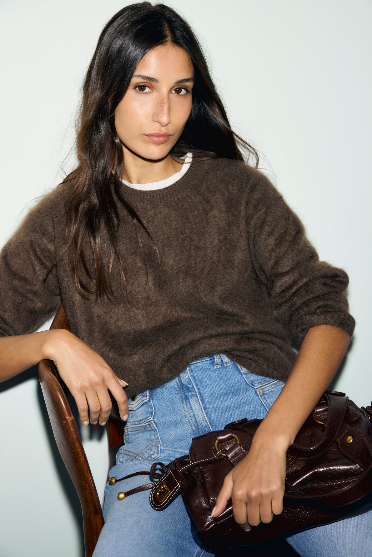 A woman with long dark hair wears a pull marron and blue jeans, sitting on a chair and holding a dark brown bag in her lap, against a plain light background.