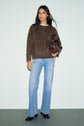 A woman stands against a plain background wearing a pull marron, light blue flared jeans, black shoes, and holding a small brown handbag.