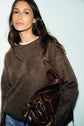 A woman with long brown hair wears a pull cachemire marron, its texture soft and luxurious, and holds a dark brown leather handbag. She stands against a plain light background, looking confidently at the camera.