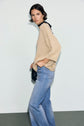 A person with short dark hair wears a beige pull lurex doré à col rond and light blue wide-leg jeans, standing sideways against a plain light background.