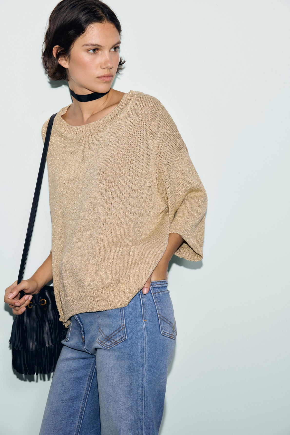 A person wearing a loose beige pull lurex doré à manches courtes, blue jeans, a black neck scarf, and carrying a black fringed bag stands against a plain light background.
