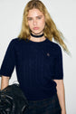 A person with long blond hair wears a bleu marine pull sans manche en laine mérinos, a black choker, and plaid pants, holding a black fringed bag against a plain white background.