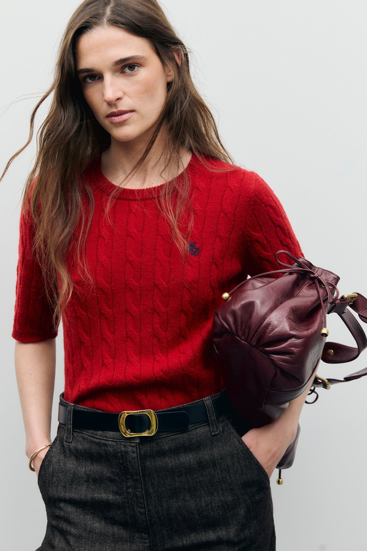 A woman with long brown hair wears a red cable-knit short-sleeve sweater crafted from soft laine mérinos, dark high-waisted pants with a black belt, and holds a burgundy leather bag under her arm, standing against a plain light gray background.