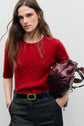 A woman with long brown hair wears a red cable-knit short-sleeve sweater crafted from soft laine mérinos, dark high-waisted pants with a black belt, and holds a burgundy leather bag under her arm, standing against a plain light gray background.
