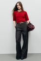 Woman stands against a plain wall wearing a red pull sans manche in laine mérinos, paired with high-waisted wide-leg dark gray trousers and black shoes, holding a burgundy handbag.