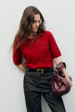 A woman in a red pull sans manche sweater crafted from soft laine mérinos and dark pants stands against a light background, holding a maroon handbag.