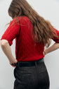 A person with long brown hair, wearing a red cable knit pull sans manche sweater and dark jeans, stands with hands on hips, facing away against a plain light background.
