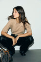 A woman with long dark hair wearing a tan knit short-sleeve top in soft laine mérinos and black pants squats against a light background. A brown and black designer handbag sits on the floor nearby.