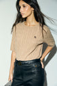 A person with long dark hair wears a tan short-sleeve knit sweater in douce laine mérinos and high-waisted black pants, standing against a light background.