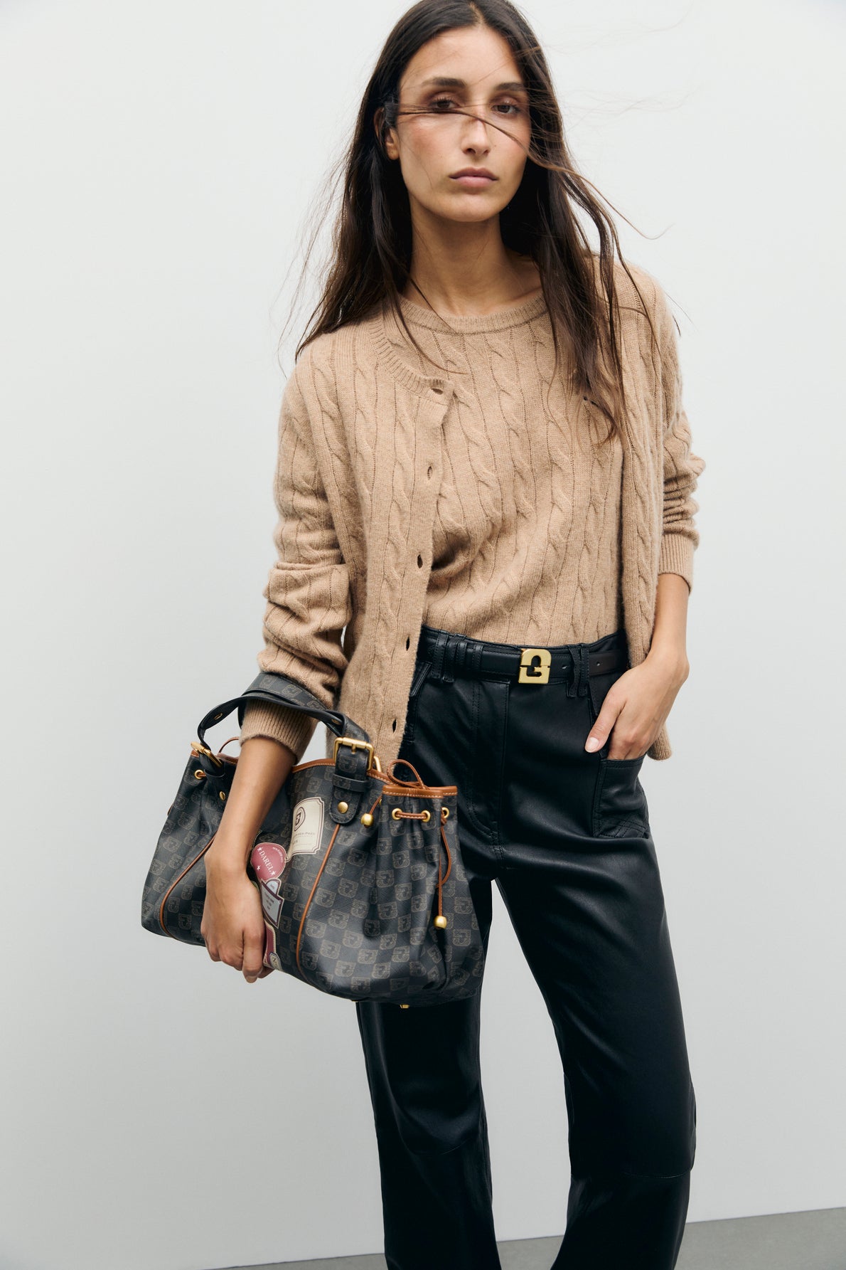 A woman wearing a tan laine mérinos cable-knit sweater and black leather pants holds a black designer handbag. She has long, loose brown hair and is standing against a plain light background.