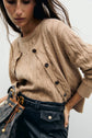 A woman wears a tan gilet à torsades crafted from soft laine mérinos with brown buttons, paired with black high-waisted leather pants featuring large pockets. She holds a drawstring bag.