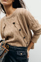 Woman wearing a light brown gilet à torsades crafted from soft laine mérinos, paired with a matching cardigan, black high-waisted pants with pockets, and holding a brown patterned handbag. Her face is partially out of frame.