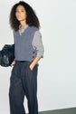 A person with curly hair wears a striped shirt, a pull col V femme gray sweater vest, and gray wide-leg pants, holding a black bag against a plain light background.