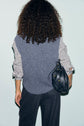 Person with curly hair, wearing a striped shirt and a pull laine sans manche gris bleuté, stands facing away. They carry a black leather bag on their left shoulder and wear dark pinstriped pants. Background is plain light blue.