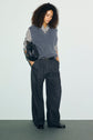 A person with curly hair wears a pull col V femme gris bleuté, styled as a sleeveless sweater over a striped shirt, wide-leg pinstripe trousers, black loafers, and carries a black handbag. They stand against a plain background, hands in pockets, looking down.