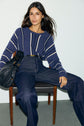 Woman with long brown hair sits on a chair wearing a pull rayé bleu jean and high-waisted wide-leg jeans, holding a textured black handbag. The background is plain and light-colored.