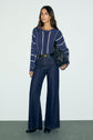 Woman standing against a plain background, wearing a stylish pull rayé bleu et blanc sweater, high-waisted wide-leg jeans, black boots, and holding a black woven handbag.