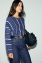 A woman with long brown hair wears a pull rayé bleu et blanc, high-waisted jeans, and a black belt while holding a textured black handbag against a plain light background.