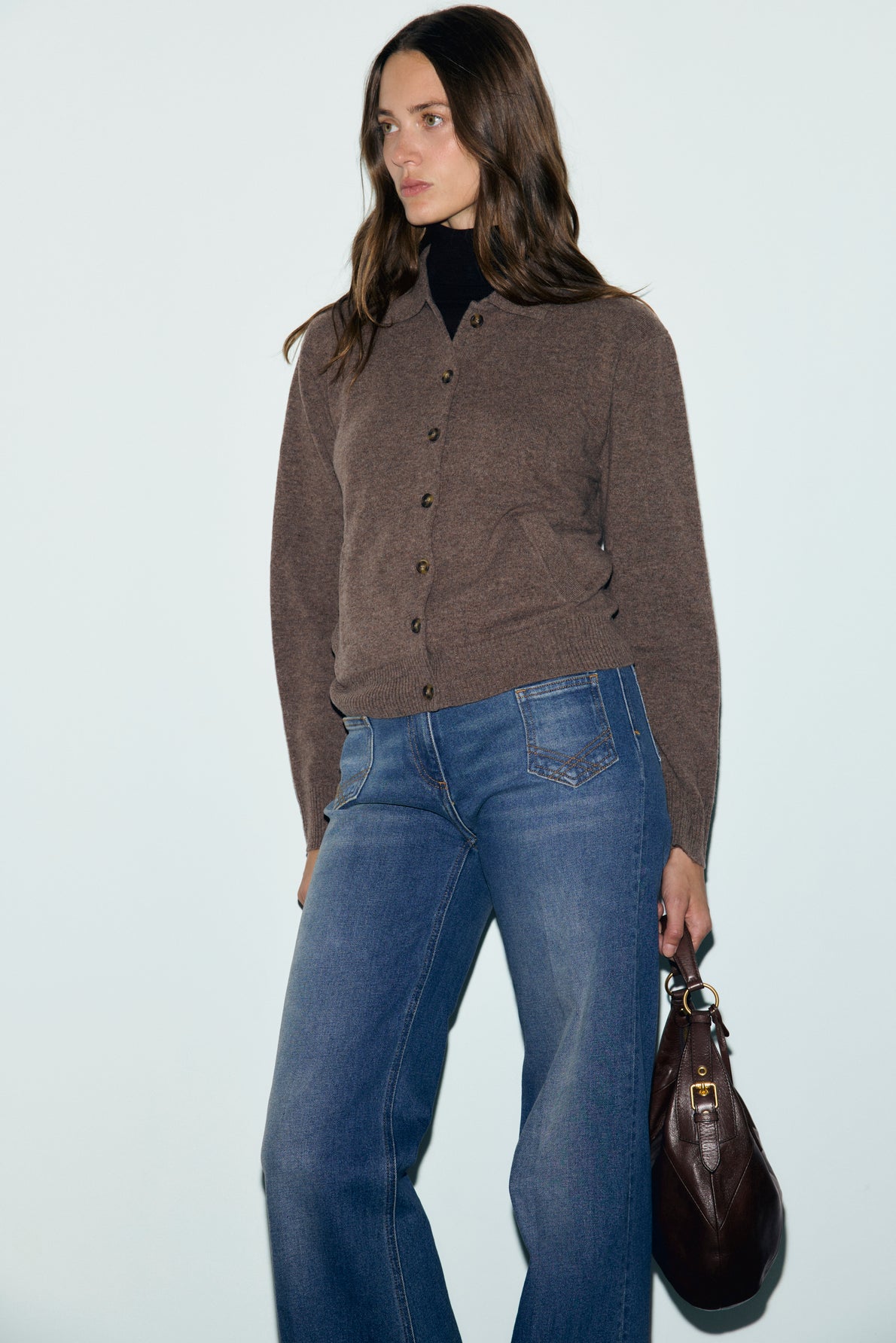 Woman with long brown hair wearing a gilet marron button-up cardigan, blue jeans, and holding a dark brown handbag, standing against a plain light background.