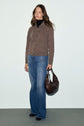 Woman stands against a plain background, wearing a gilet marron over a black turtleneck, blue jeans, and black shoes. She holds a dark brown handbag in her left hand and has long, wavy hair.