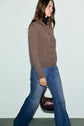 A woman with long brown hair walks sideways, wearing a gilet marron button-up sweater, blue jeans, and holding a dark brown handbag. She is in front of a plain light background.