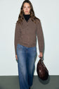Woman with long brown hair wears a cardigan laine in brown, paired with a black turtleneck, wide-leg jeans, and holds a large dark brown bag, standing against a plain light gray background.