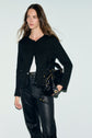 A woman with long brown hair wears a black textured gilet noir jacket with subtle lurex sequins, a white top, and black leather pants. She holds a black handbag and stands against a plain light background.