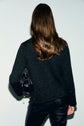 A person with long brown hair, wearing a textured black sweater layered with a gilet court and black pants, stands facing away from the camera, holding a small black handbag.