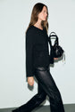 A woman with long brown hair walks while holding a small black handbag. She is wearing a textured black sweater layered under a stylish gilet noir and black leather pants against a plain light background.